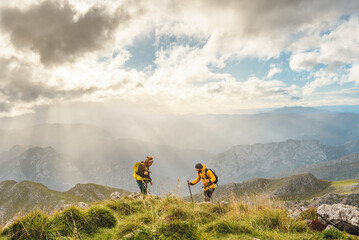 Obraz premium couple of hikers, man and woman, reaching the top of a mountain on a day with clouds and sun. Two mountaineers hiking equipped with backpacks and trekking poles. mountainous landscape from height.