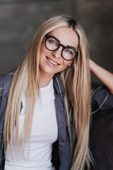 Vertical portrait of gorgeous blonde Swedish woman in glasses, grey shirt with long loose hair leaning on hand looks at camera toothy smiles, satisfied by career of psychologist. Model posing indoors.