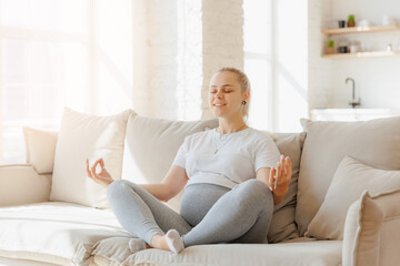 Concept meditating health pregnancy. Portrait beautiful young pregnant woman practicing yoga at home on sofa, background light kitchen