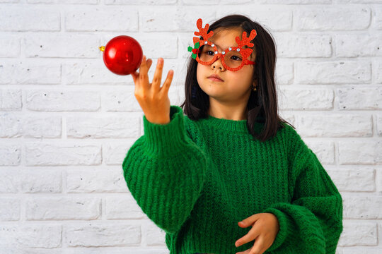 Asian girl in masquerade glasses with reindeer horns, playing with a Christmas ball