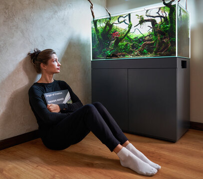 A beautiful woman sitting on the floor with the book in the hands and looking to the aquarium aquascape.