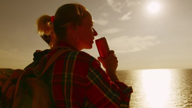 Traveler woman friends drinking drink from tin can lit by sunset golden sun top of cliff overlooking sea.