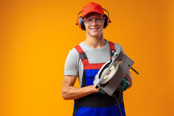Young male construction worker with modern circular saw on yellow studio background