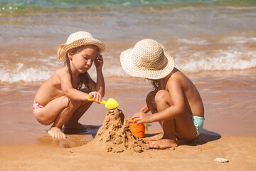 Two little girls sisters in swimsuits and sun hat playing with sand on tropical sandy beach at...