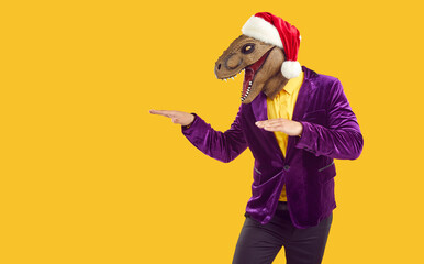 Portrait young man wearing Christmas cap and funny excited dinosaur reptile lizard face masquerade mask advertising monster discounts and showing creative copy space side on yellow studio background