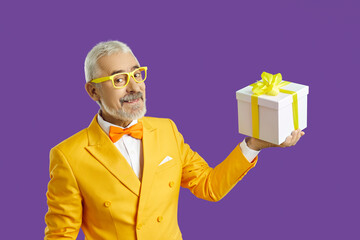 Studioportret van een volwassen man die aanwezig is. Gelukkig knappe senior man in fel geel funky stijlvol pak en bril met mooie witte geschenkdoos met geel lint. Vakantie verkoopconcept
