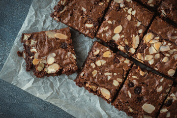 Delicious homemade chocolate brownies with almond flakes and raisins cut on square slices on the baking parchment. Flat lay. Grey concrete background