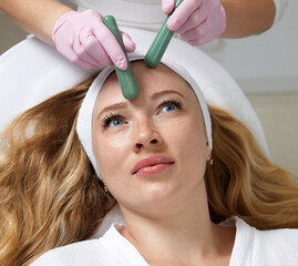 Fototapeta premium Young woman having facial massage with stone sticks in spa salon