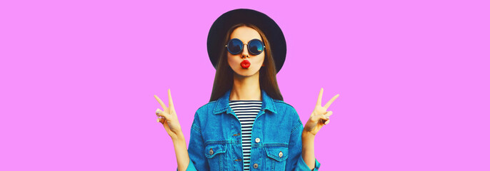Portrait of stylish young woman blowing her red lips sending air kiss wearing black round hat, jean jacket on pink background, blank copy space for advertising text