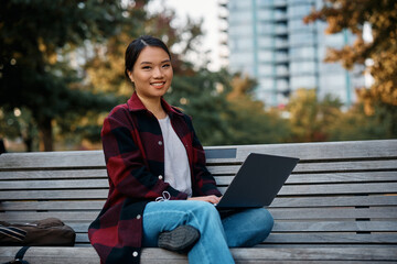 Happy Asian woman using laptop in park and looking at camera.