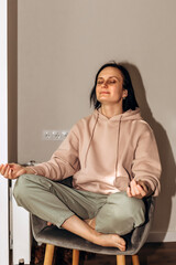 Fototapeta premium A thoughtful girl in a pink hoodie sits on a gray chair near the window and radiator with closed eyes and doing meditation. Front view