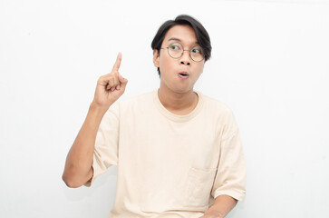 asian young man in casual beige tshirt pointing finger showing something good. man with glasses shocked happily pointing up with copy space isolated over white background. advertisment model concept.