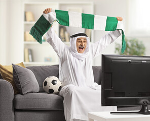 Excited arab man in a traditional robe holding a scarf and watching football on tv