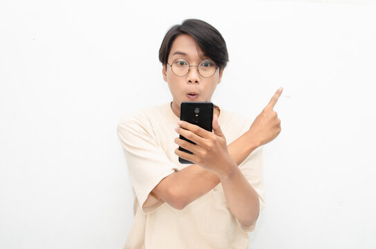 excited young asian man in beige tshirt holding and showing phone screen with happy euphoric shocked wow expression. man pointing to screen phone with thumb up. billboard model advertisment concept.