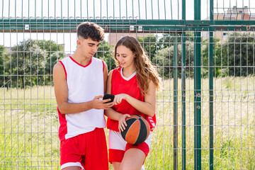 Young man and woman basketball players using phone together in park