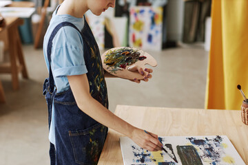 Cropped shot of young craftswoman with color palette working over painting by workplace while using...