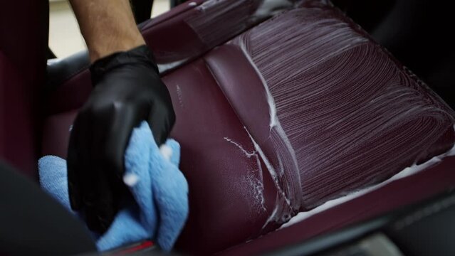 Professional cleaner in gloves carefully dry cleaning the burgundy leather car seat with the rag and foam close up. Intensive car interior manual dry cleaning