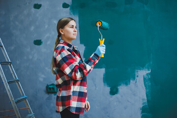 Fototapeta premium A young beautiful woman designer makes repairs, paints a gray wall with green paint with a roller. Plaid shirt, long hair and jeans.