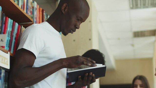 African male college student standing and reading a book in library with lots of books. University student looking for study references.