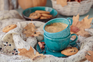 Autumnal composition with white knitted scarf, blue cup of coffee, cookies and dry yellow leaves on a table. Autumn mood.
