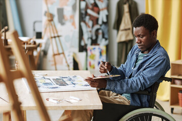Young African American craftsman in wheelchair mixing colors on palette while sitting by table in...