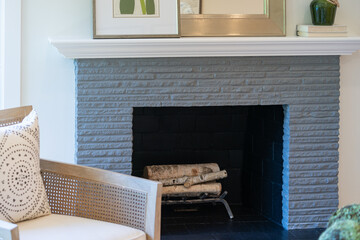 Old fashioned painted brick fireplace with birch logs, white wood mantlepiece and neutral rattan cushioned side chair.