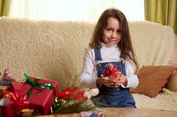 Lovely little child girl with long hair, sitting on sofa at cozy home interior with Christmas gift box in her hands, cutely smiles looking at camera. Winter holidays atmosphere. New Years preparations