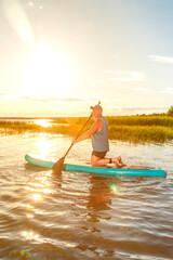 a man in shorts and a T-shirt on a SUP board with an oar floats on the water against the background of the setting sun.