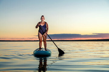 a woman in a closed swimsuit with a mohawk standing on a SUP board with an oar floats on the water against the background of the sunset sky.