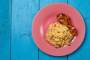 Couscous with vegetables in sauce and spices with grilled chicken drumstick on a pink plate on a blue table.
