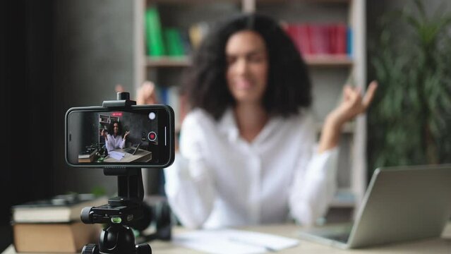 Blurred relaxed young woman in casual wear sitting on workplace with modern laptop and meditating with closed eyes during recording video blog. Focus on modern smartphone fixed on tripod.