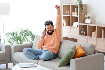 Happy young bearded man talking by mobile phone on grey sofa at home