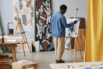 Back of young black man in casualwear working over new painting while standing in front of easel...