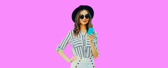 Portrait of stylish happy smiling young woman model with smartphone wearing black round hat, striped shirt on pink background, blank copy space for advertising text