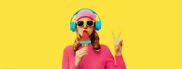 Summer fresh colorful portrait of stylish woman in headphones listening to music with fruit juicy lollipop or ice cream shaped slice of watermelon on yellow background - obrazy, fototapety, plakaty