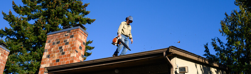 Senior man with gas powered leaf blower cleaning roof gutters on an apartment building, fall maintenance
