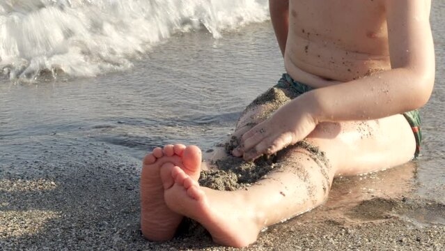 kid child boy having fun on sands beach seashore digging throwing wet sand on legs splashing hitting with hands the water.sea waves sun lith sunshine,vacation family.orange rake toy in focus 4k video.