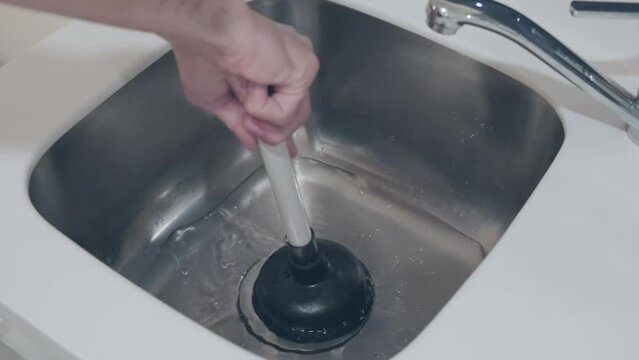 Hands hold plunger with wooden handle for pipe cleaning against the backdrop of kitchen sink. Drain clog cleaning work. Repairman, plumber, cleaning service man at work.