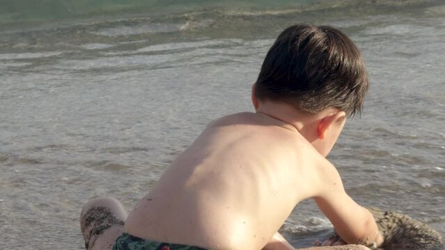 kid child boy having fun on sands beach seashore digging throwing wet sand on legs splashing hitting with hands the water.sea waves sun lith sunshine,vacation family.orange rake toy in focus 4k video.