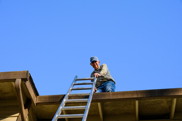 Naklejka na ściany i meble Senior man on apartment building roof ready to climb down an aluminum extension ladder 