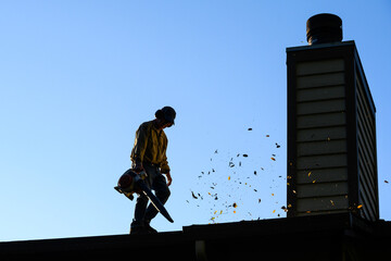 Silhouette of senior man with gas powered leaf blower cleaning roof gutters on an apartment...