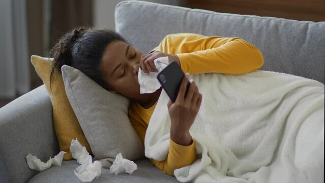 Feeling unwell. Young sick african american woman lying on sofa at home, browsing on phone and blowing nose into tissue