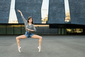 Woman in street clothes and ballerina shoes dancing outdoors on concrete.