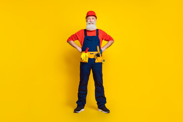 Full length photo of positive confident age man wear overall uniform red hard hat arms waist isolated yellow color background