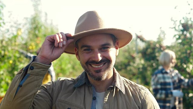 Close up of happy young Caucasian handsome man in hat standing in green apple orchard and smiling to camera. Outside. Harvesting concept. Portrait of attractive male farmer at fruit crop gathering.