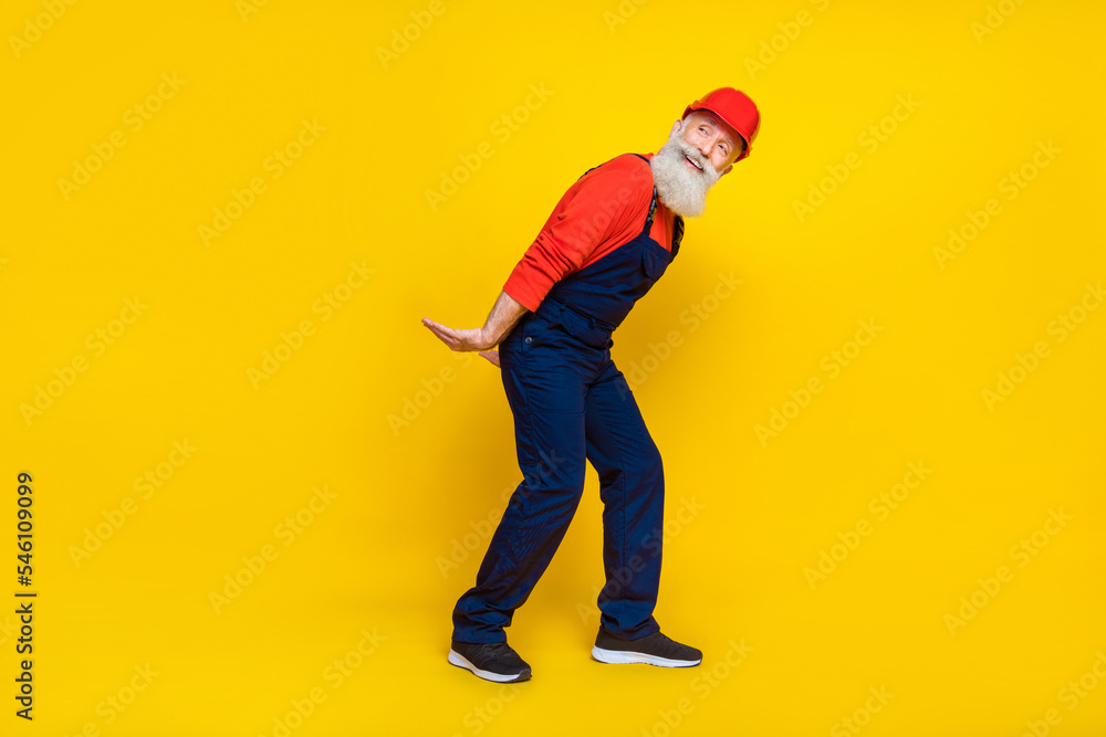 Wall mural Photo of funky cheerful senior guy dressed uniform overall red hardhat holding arms heavy empty space isolated yellow color background