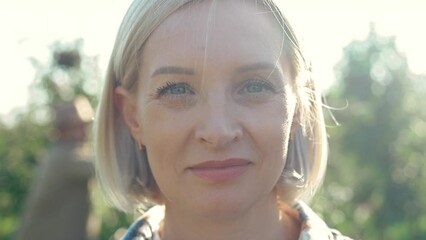Close up of face of young Caucasian beautiful blonde woman smiling and looking straight to camera in apple orchard. Outdoors. Portrait shot of female gardener among green trees. Harvest concept.