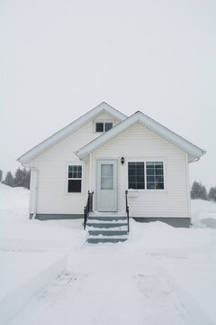 Light snow falling on a cozy small house in a rural setting with a light sky and deep snow on the ground