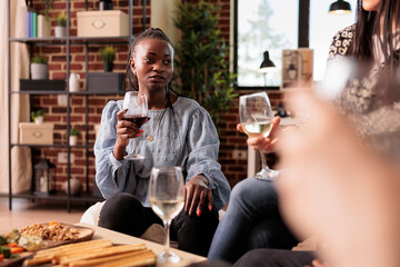 African american lady drinking wine, listening, hearing attentively friends conversation, talking, discussion, chatting at home, house, apartment party, gathering, reunion.