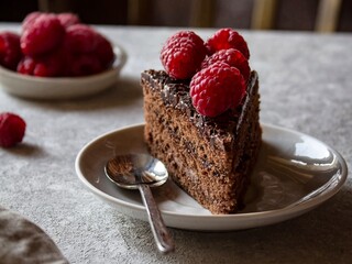 A piece of delicious homemade chocolate cake on a plate, decorated with raspberries on top. Chocolate dessert and raspberries on the table.